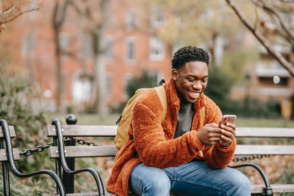 picture of young man smiling at phone sitting on the bench in the park young african man