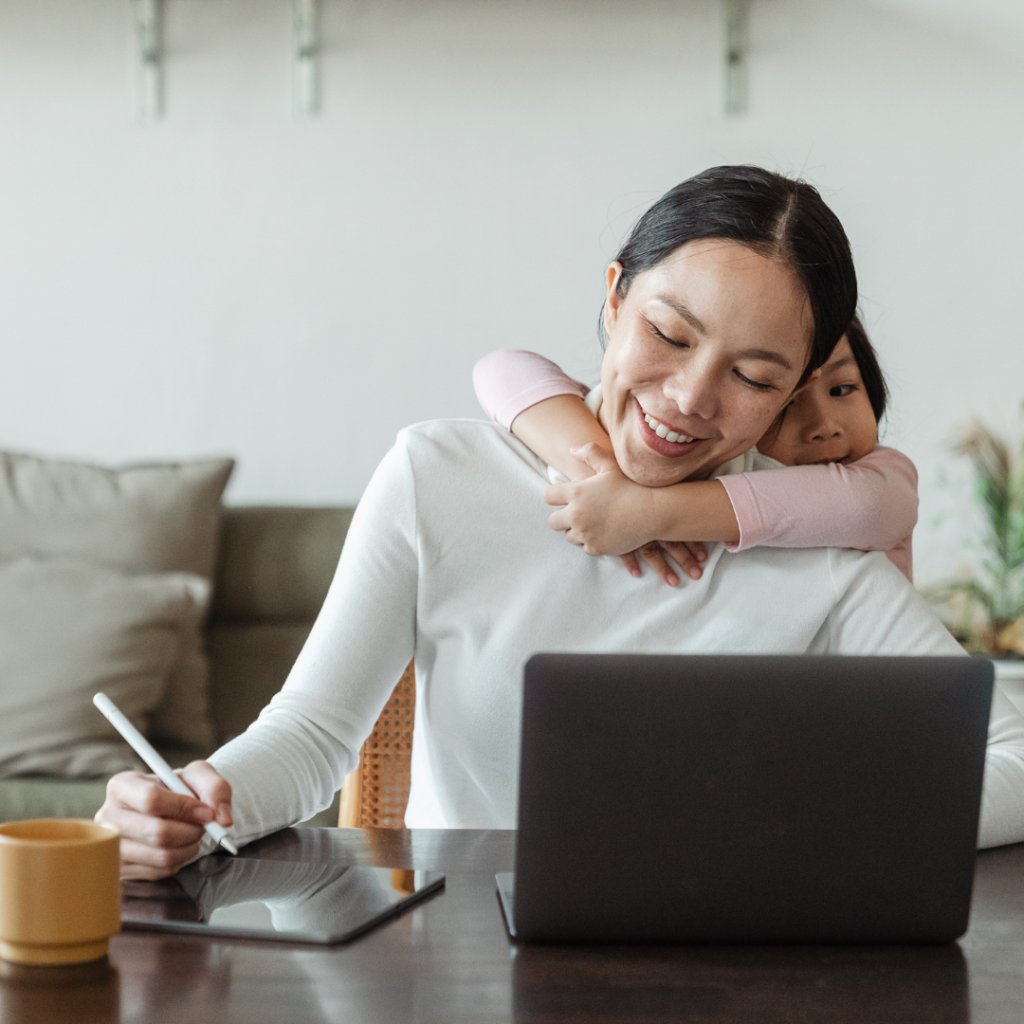 Mum sitting at desk working on laptop with child hugging from behind, remote work