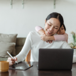 Mum sitting at desk working on laptop with child hugging from behind, remote work