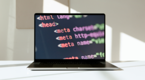 A laptop on a white surface with its screen displaying lines of HTML code. The code includes tags such as , , , and other meta elements